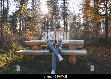 Musicien assis sur un banc en bois rustique dans une forêt ensoleillée, jouant du ukulélé. Entouré par le feuillage d'automne et la lumière chaude du soleil coulant à travers le t Banque D'Images