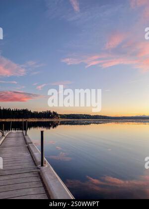 Un quai flottant s'étend dans un lac tranquille pendant le coucher du soleil, reflétant des teintes roses et oranges vibrantes dans l'eau sereine, avec un horizon boisé und Banque D'Images