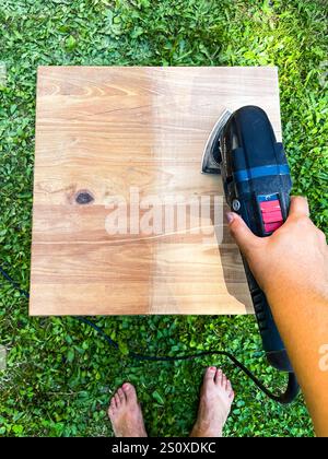 Main de femme ponçant vieux tabouret en bois par outil électrique sur l'herbe verte dans le jardin Banque D'Images