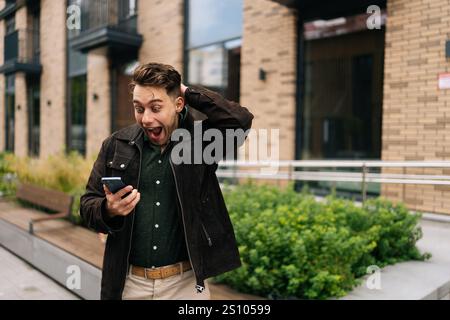 Heureux jeune homme excité debout dans la rue de la ville, tenant un téléphone portable et réagissant avec étonnement à certaines nouvelles inattendues qui viennent d'être reçues. Banque D'Images