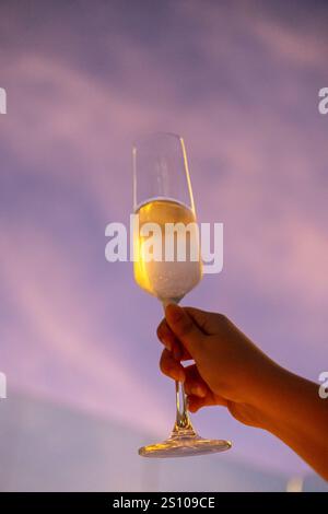 Une main tenant un verre de Prosecco ou de vin mousseux avec un ciel de coucher de soleil coloré sur le fond Banque D'Images