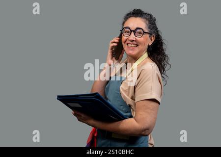 Femme joyeuse portant tablier et lunettes, parlant sur smartphone et tenant dossier, portrait de studio sur fond gris Banque D'Images