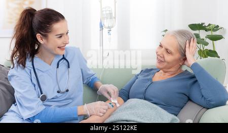 La jeune infirmière souriante ou le médecin aide la patiente âgée malade âgée à recevoir l'infusion goutte à goutte IV et la thérapie d'injection de vitamine assis sur le canapé en moi Banque D'Images