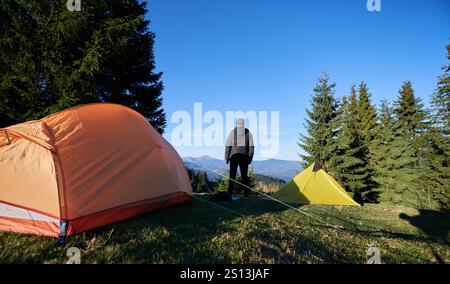 Vue arrière du camping-car se dresse entre la tente orange et jaune, regardant les montagnes lointaines sous le ciel bleu. Grands arbres à feuilles persistantes, ombres projetées sur un camping herbeux. La lumière du matin illumine le paysage. Banque D'Images