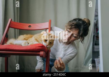 Jeune femme joue avec son chat gingembre sur une chaise rouge, créant une scène réconfortante d'amour et d'affection dans un cadre confortable à la maison Banque D'Images