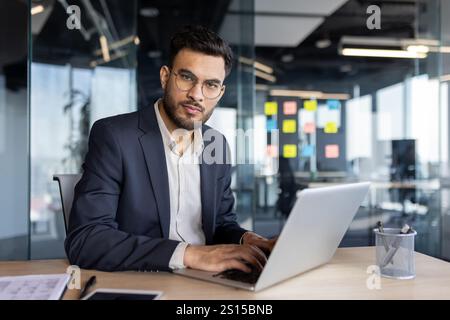 Homme d'affaires latino-américain confiant utilisant un ordinateur portable assis dans un environnement de bureau contemporain. Professionnel ciblé mettant en valeur la productivité et la détermination dans un espace de travail. Banque D'Images