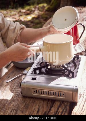 Main ouvrant le couvercle d'une casserole placée sur une cuisinière à gaz portable sur une table en bois, cuisiner à l'extérieur Banque D'Images