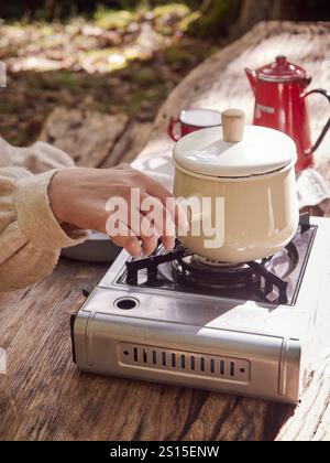 Main tenant la poignée du pot sur la cuisinière portative à gaz sur une table en bois, cuisson à l'extérieur Banque D'Images