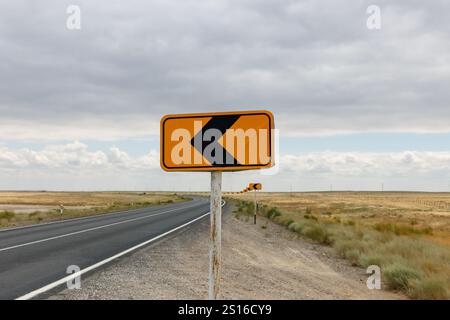 Un panneau de virage à gauche jaune vif se dresse à côté d'une route sinueuse au Kazakhstan, avec un vaste paysage ouvert qui s'étend au-delà. Banque D'Images