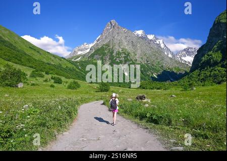 Une jeune fille touriste avec un sac à dos marche le long d'un chemin de terre parmi les montagnes avec des sommets couverts de neige. Montagnes Dombay, Russie Banque D'Images