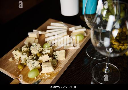 Un plateau de fromage en bois servi avec divers fromages, raisins et olives, magnifiquement arrangé, accompagné de verres de vin, créant une ambiance élégante Banque D'Images