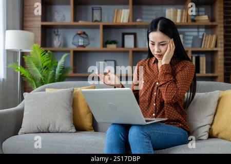 Femme asiatique se sentant stressée tout en travaillant sur un ordinateur portable à la maison, assise sur un canapé confortable entouré de coussins et de plantes. Une expression incertaine indique des difficultés liées au travail à domicile. Banque D'Images