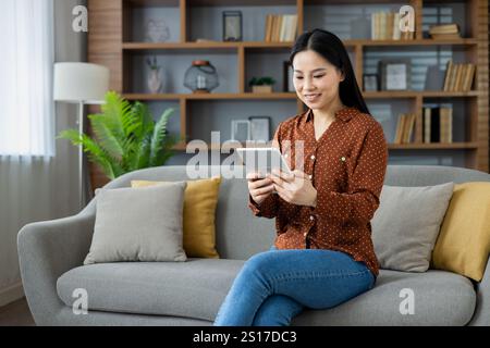 Jeune femme asiatique assise à la maison sur le canapé souriant joyeusement en utilisant une tablette, en utilisant une application éducative et en lisant des livres en ligne, en regardant des flux vidéo. Banque D'Images