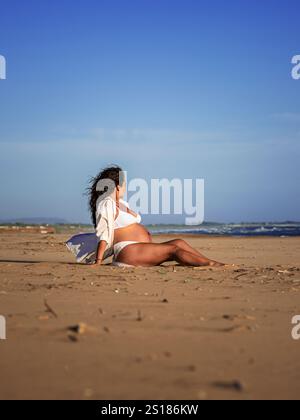 belle femme méditerranéenne aux cheveux bouclés enceinte pose sur la plage Banque D'Images