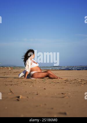 belle femme méditerranéenne aux cheveux bouclés enceinte pose sur la plage Banque D'Images