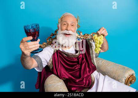 Portrait de l'ancien chef d'homme riche assis trône soulever le raisin de verre de vin isolé sur fond de couleur bleue Banque D'Images