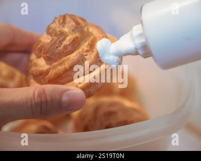 La main féminine tient une pâte à choux fantaisie ronde ou eclair et la remplit de crème anglaise à l'aide d'une seringue de confiserie. Banque D'Images