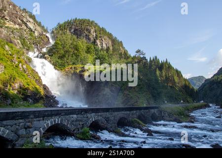 Cascade coule de façon spectaculaire sur des formations rocheuses dans une rivière en contrebas, entourée d'arbres et de collines vibrants. Un pont de pierre enjambe la rivière. Banque D'Images