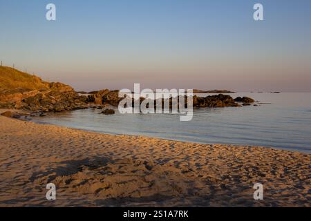 De douces vagues se battent contre la rive sablonneuse de Farsund, en Norvège, tandis que le soleil se couche sur une plage tranquille. Des rochers émergent de l'eau, entourés de teintes douces Banque D'Images
