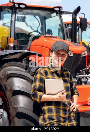 Revendeur de matériel agricole ou de construction. Femme avec table numérique debout à côté du tracteur de couleur orange à la concession. Banque D'Images