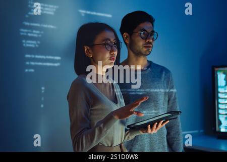 Asiatique deux développeurs de logiciels engagés dans une session de codage collaboratif, avec du code projeté sur le mur et plusieurs écrans d'ordinateur s'affichant Banque D'Images