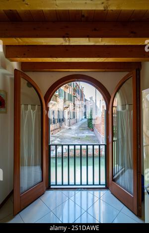 Vue à travers les doubles portes voûtées donnant sur un étroit canal vénitien et des bâtiments rustiques. L'intérieur présente des poutres en bois et des sols carrelés, blendi Banque D'Images