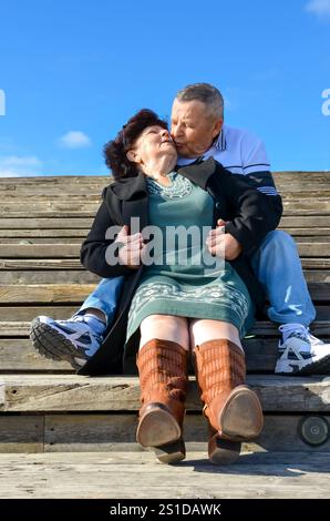 Couple senior profitant d'un moment romantique, embrassant sur des marches en bois extérieures sous un ciel bleu clair Banque D'Images