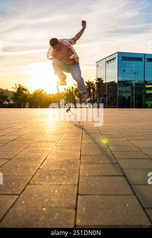 Beau jeune patineur faisant un tour acrobatique au coucher du soleil - homme cool moderne portant des vêtements urbains élégants se promenant dans la ville avec du skateboard Banque D'Images