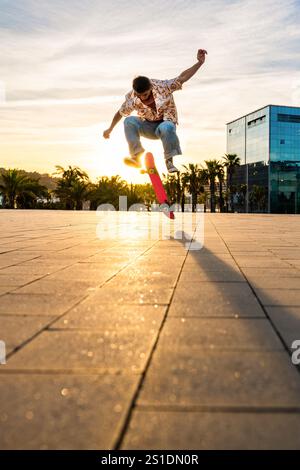 Beau jeune patineur faisant un tour acrobatique au coucher du soleil - homme cool moderne portant des vêtements urbains élégants se promenant dans la ville avec du skateboard Banque D'Images