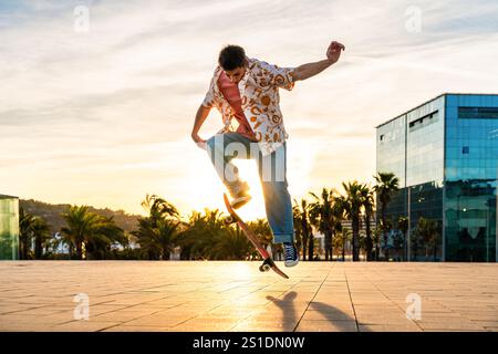 Beau jeune patineur faisant un tour acrobatique au coucher du soleil - homme cool moderne portant des vêtements urbains élégants se promenant dans la ville avec du skateboard Banque D'Images