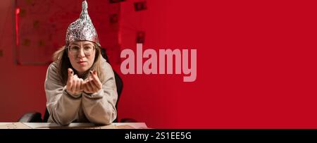 Femme portant des lunettes dans un chapeau en aluminium montrant Give me Money geste avec les doigts assis sur fond rouge. Complot. Théorie du complot. Bannière. Copier sp Banque D'Images