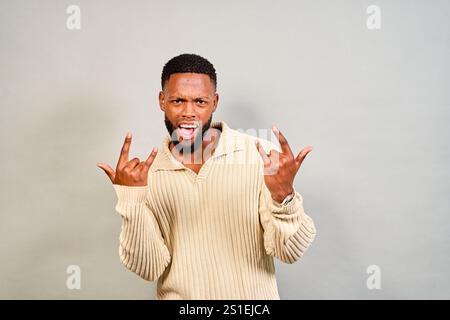 Le jeune homme exprime énergiquement son amour pour le rock and roll, en faisant un symbole de main et en criant avec excitation sur un fond gris. Son look tendance c Banque D'Images