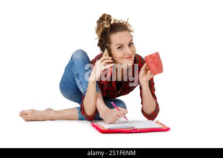 Jeune femme prend des notes dans un cahier, boit du café et parle sur un téléphone portable assis dans une pose de yoga isolée sur un fond blanc Banque D'Images