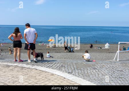 Aberystwyth Royaume-Uni - 2 août 2024 ; couple avec petit chien debout sur le chemin de la plage et le front de mer avec les vacanciers et les bains de soleil appréciant Banque D'Images