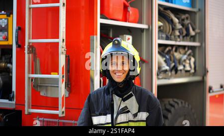 Portrait d'heureux garde-feu féminin sur fond de grand camion rouge. Jeune femme de feu en équipement complet regardant la caméra avec des émotions positives Banque D'Images