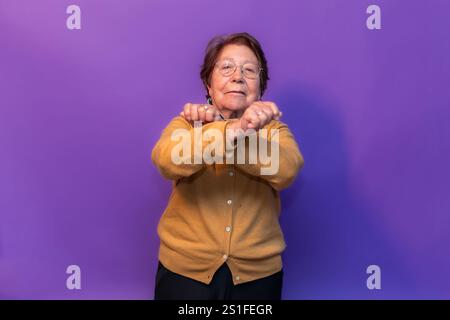 Femme âgée déterminée croisant les bras et signalant un arrêt avec ses mains sur un fond violet vibrant, incarnant la résilience et l'autonomisation des hommes Banque D'Images