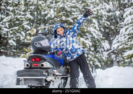 Petit garçon sur la motoneige dans les montagnes souriant par temps neigeux, forêt. Enfant monte une motoneige bleue sur un sentier enneigé, pins. Garçon Banque D'Images