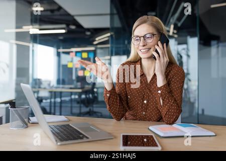 Femme d'affaires joyeuse et heureuse parlant au téléphone tout en étant assise au bureau à l'intérieur du bureau. Le travailleur de bureau est satisfait de travailler avec un ordinateur portable et des documents. Banque D'Images