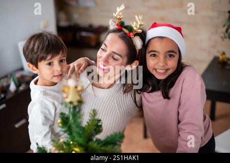 Une femme tient un petit sapin de Noël avec ses deux enfants. Les enfants portent des chapeaux de Père Noël et la femme porte un bandeau de cerf. Scène i. Banque D'Images