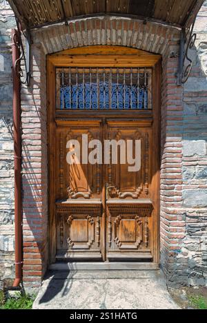 Une porte en bois magnifiquement conçue présente des sculptures complexes, encadrées par un bâtiment en pierre. La lumière du soleil met en valeur les textures, créant des ombres qui rehaussent les détails et le charme de la porte Banque D'Images
