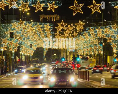 Athènes, Grèce - 3 janvier 2025 : rue urbaine illuminée par des lumières en forme d'étoile dorée et la circulation pendant la saison des fêtes. Acropole en vue. Banque D'Images