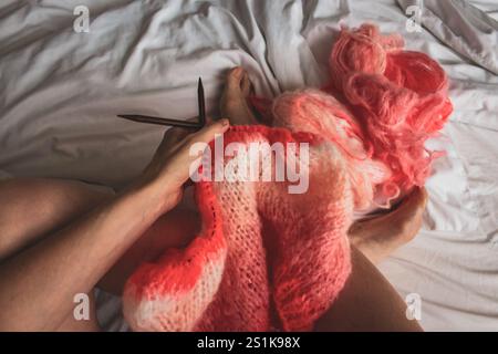 Femme avec fil de corail et aiguilles à tricoter en bois sur drap de lit blanc, vue de dessus. Jambes pour femmes et pull tricoté brillant, vue d'en haut. Passe-temps de tricot Banque D'Images