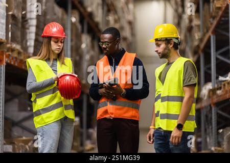 Une équipe d'employés de l'entrepôt discute des procédures de sécurité tout en portant des casques et des gilets réfléchissants. Ils sont debout dans une allée entourée de shel Banque D'Images