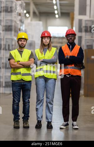 Un groupe diversifié d'ouvriers d'entrepôt portant des gilets de sécurité et des casques, debout avec confiance dans une grande zone de stockage. L'image reflète le travail d'équipe, la plongée Banque D'Images