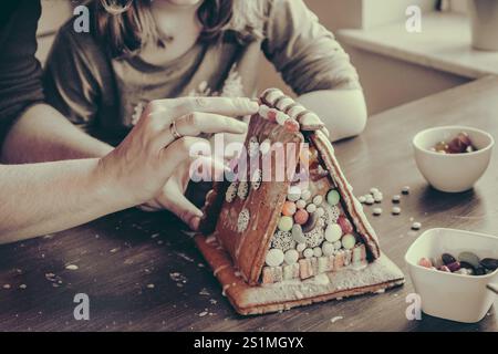 Enfant et femme décorent une maison en pain d'épice avec des bonbons colorés, style vintage désaturé Banque D'Images