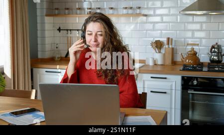 Une femme aux longs cheveux bouclés est assise dans une cuisine, portant un casque et engageant une conversation avec un appel vidéo en ligne ou une session de service client Whil Banque D'Images
