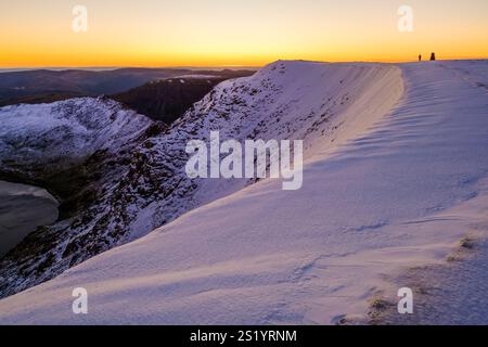 Un marcheur arrive au pilier trig sur Helvellyn dans le Lake District quelques minutes avant le lever du soleil par un matin frais d'hiver. La lueur orange du Banque D'Images