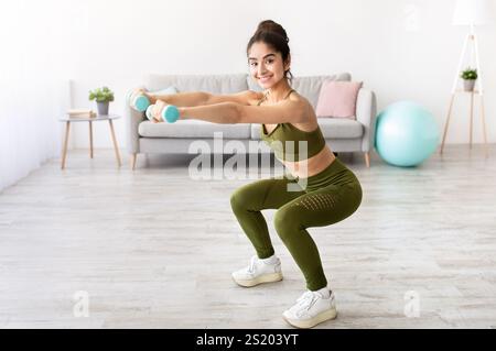 Jeune femme indienne forte qui travaille avec des haltères, qui s'accroupite à la maison, dans un espace vide Banque D'Images