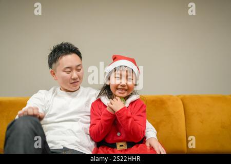 Un jeune parent et enfant japonais, une fille de 6 ans et un homme dans la vingtaine, sont assis sur un canapé à Noël, habillés en Père Noël. Banque D'Images