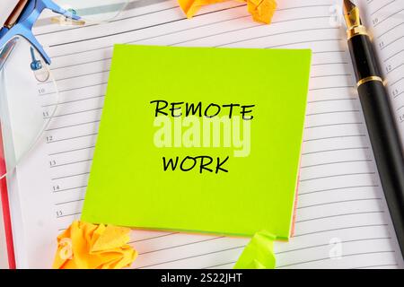 Note collante brillante inscrite avec travail à distance placé sur un bloc-notes entouré de papiers froissés et d'instruments d'écriture sur un espace de travail. Banque D'Images